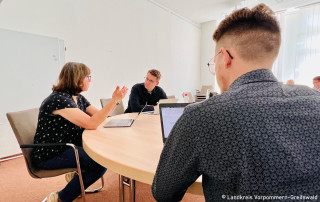 Personen die zusammen mit ihren Laptops an einem Tisch sitzen und sich austuaschen.