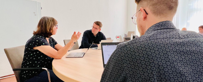 Personen die zusammen mit ihren Laptops an einem Tisch sitzen und sich austuaschen.